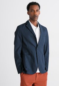 Sacou bleumarin închis, realizat dintr-un material texturat, cu un design cu un singur nasture, guler în formă de V, buzunare frontale și o croială slim, asortat cu o cămașă albă.