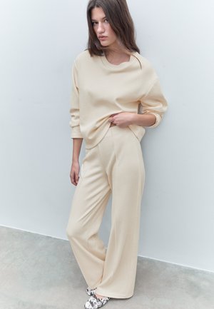 Jeune femme portant un pull beige ample et un pantalon large, debout, une main tenant son pull, devant un mur clair uni.
