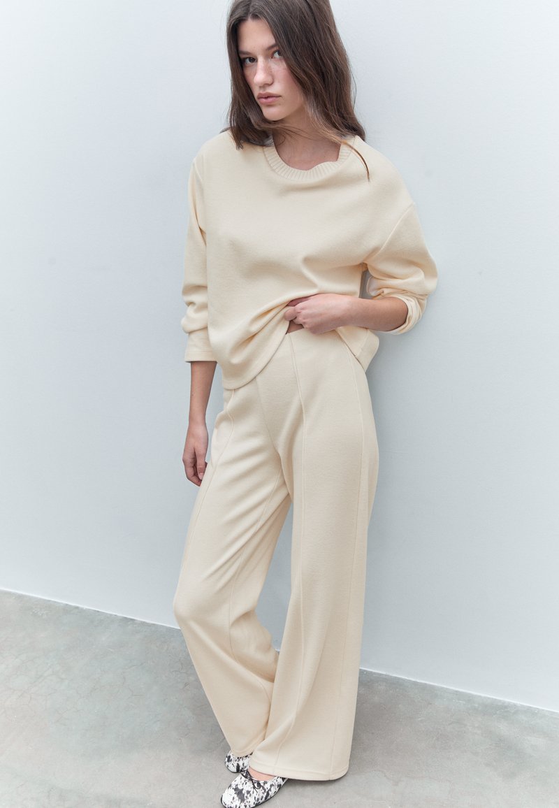 Giovane donna con un maglione beige largo e pantaloni a gamba larga, in piedi con una mano che stringe il maglione, davanti a una parete chiara e semplice.