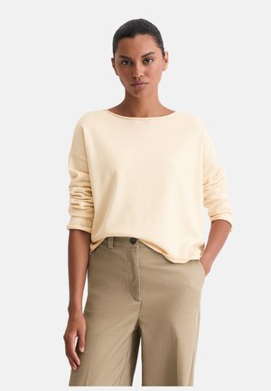 Pull long à manches longues de couleur beige clair avec un large décolleté et des poignets retroussés, associé à un pantalon kaki. Texture douce et coupe décontractée.
