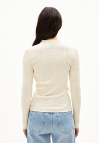 Langærmet, ribbestrikket top i off-white, med høj hals og tætsiddende silhuet, parret med lyseblå højtaljede jeans.