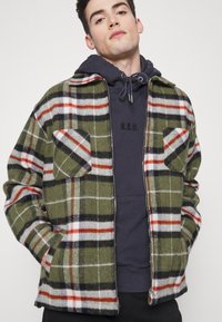 Casaco xadrez verde com accents em vermelho e preto, apresentando dois bolsos frontais, um fecho éclair e um tecido suave e texturizado. Usado sobre um moletom azul marinho.