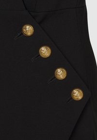 Zwarte stoffen jurk met een diagonaal ontwerp, vier gouden knopen en decoratieve stiksels aan één kant.