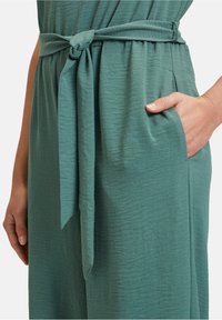 Grünes, strukturiertes Kleid mit gebundenem Taillengürtel und seitlicher Tasche. Der Stoff wirkt weich und leicht mit einem dezenten Glanz.