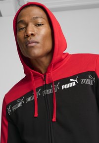 Puma zip-up kapuca, ki ima črno telo in rdečo kapuco. Opazno belo logotipno tiskanje čez prsi in rdeče vrvice za zategovanje. Gladka tekstura tkanine.