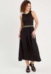 Robe maxi noire avec texture côtelée, design sans manches et taille froncée mettant en avant une ceinture décorative à rayures dorées. Associée à des sandales noires.