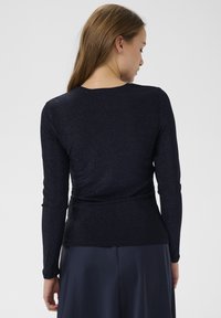 Dlhý rukáv, tmavomodrá blúzka vyrobená z mierne lesklého materiálu. Priliehavý strih s jemným zberaním v oblasti pásu a okrúhlym výstrihom.