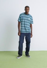 Young man standing wearing a blue striped T-shirt, dark pants, and dark sneakers on a light green floor with a plain background.