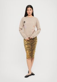Maglione beige a maglia con dettagli ricamati, abbinato a una gonna a tubino gialla a strisce di tigre. Scarpe nere flat completano l'outfit.