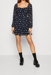 Marinblå blommig klänning med fyrkantig halsringning, puffade långärmar och volangkant. Kombinerad med svarta platåskor och strumpor.