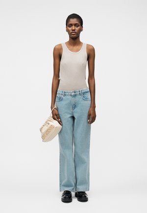 Naine kannab beežikat varrukateta tippu ja helehalli laia säärega pükse, hoides käes beežikat volditud käekotti ning kandmas musti kingi, seisab sirgelt.