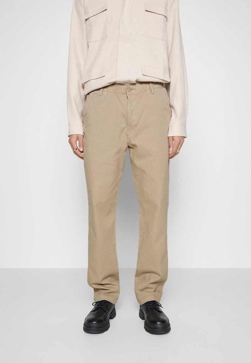 Pantalons en coton beige à coupe décontractée, avec une taille élastique, des poches latérales, portés avec des chaussures noires et une chemise de couleur claire.