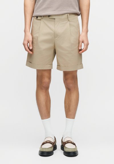 Pantalon court beige avec revers roulés, présentant des plis à l'avant. Porté avec des chaussettes blanches et des mocassins bicolores.