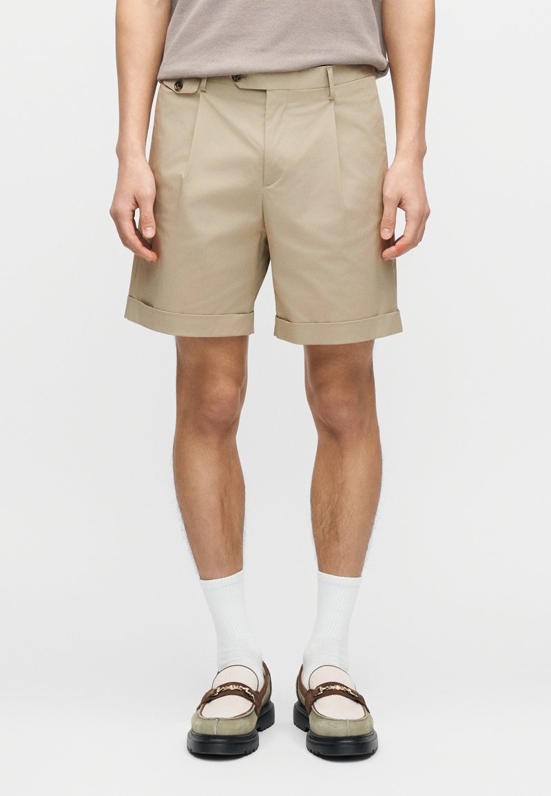 Μπεζ κοντό παντελόνι με γυρισμένα  μανίκια, με πιέτες μπροστά. Φοριέται με λευκές κάλτσες και δίχρωμα μοκασίνια.