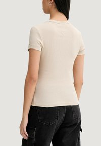 Béžový krátký top s krátkými rukávy a žebrovanou texturou, s přiléhavým střihem, širokým výstřihem a jemnými šicími detaily, nošený s černými cargo kalhotami.