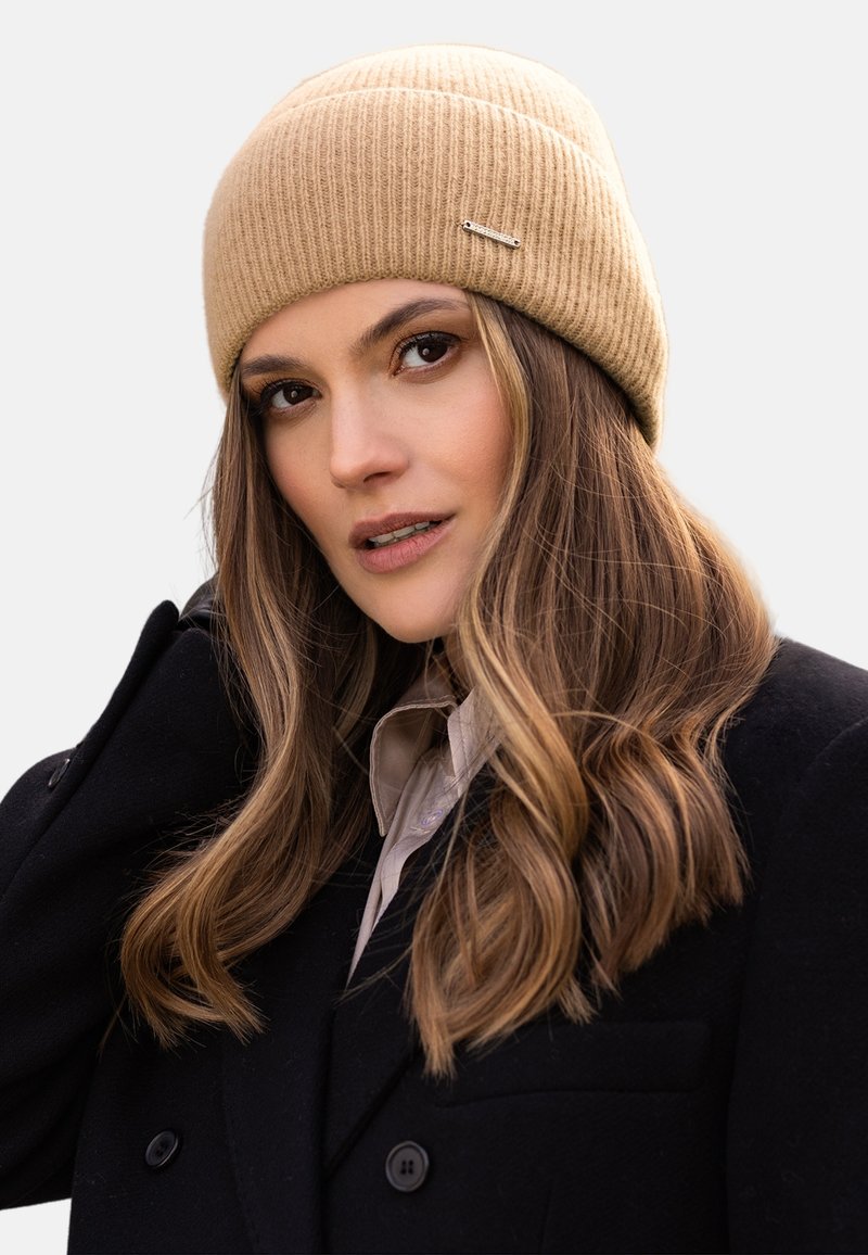 Beige geribbelde gebreide beanie met een omgeslagen ontwerp, voorzien van een kleine zilveren hardware-accent, gedragen met een zwarte jas en een licht overhemd.