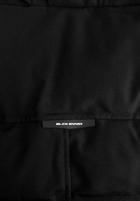 Zwarte stoffen tas met een gladde textuur en een horizontaal logo etiket dat op de voorkant is gestikt. Het ontwerp is eenvoudig en functioneel.