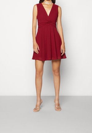 Kvinde iført ærmeløs dyb rød V-hals kjole med samlet talje, klare remsandelhøjhæle og en perlehalskæde, stående mod hvid baggrund.