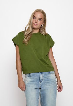Femme blonde portant un haut vert à manches courtes et un jean bleu clair, debout devant un fond blanc uni.