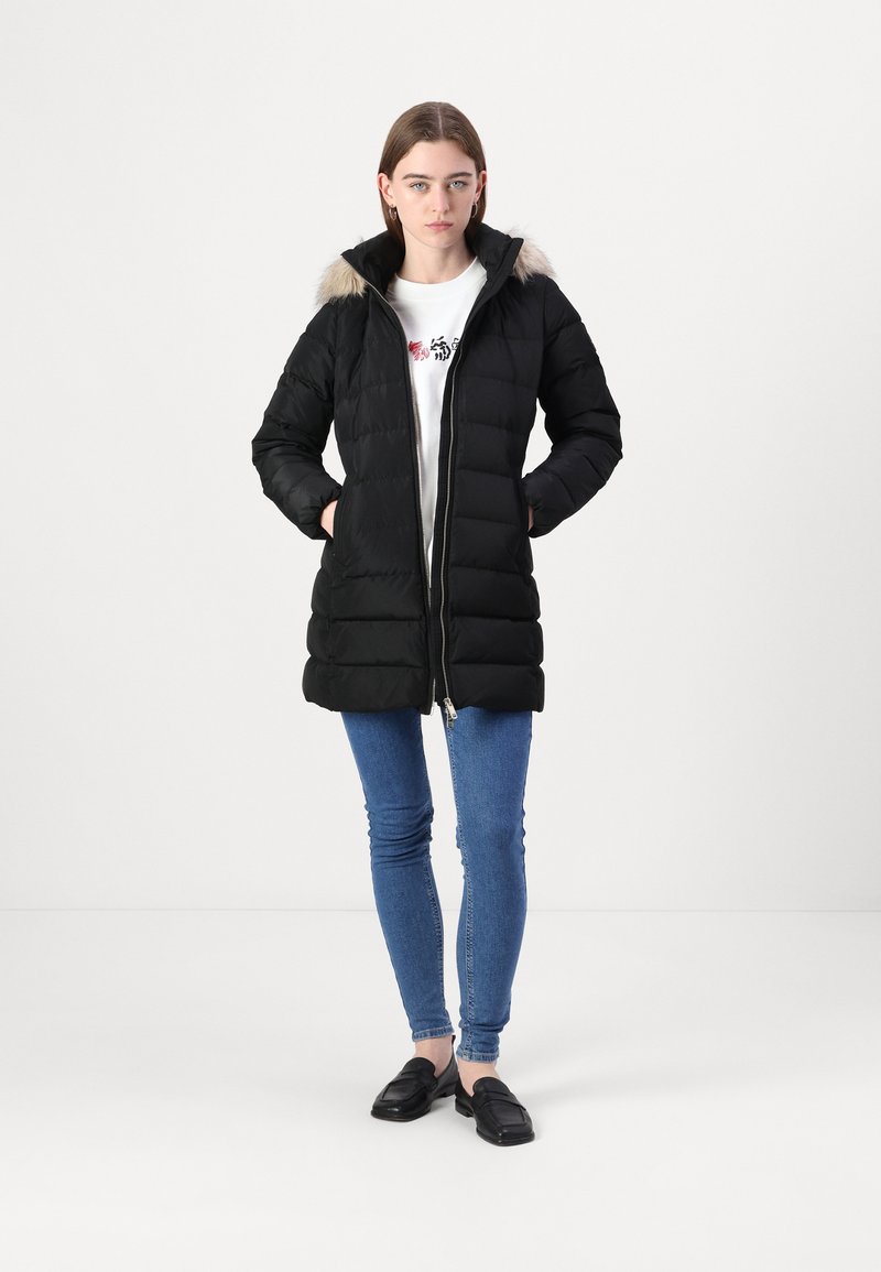 Zwarte pufferjas met een capuchon van bont, ritssluiting en een gestikt ontwerp. Gecombineerd met een wit shirt, blauwe jeans en zwarte instappers.