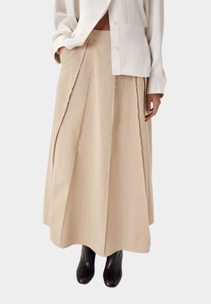 Mujer con falda beige en línea A con costuras de borde sin rematar, camisa blanca abotonada y botines negros, de pie con una mano en el bolsillo.