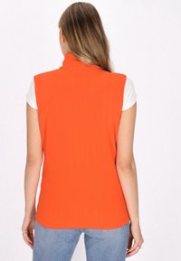 Orange ärmelloses Oberteil mit hohem Kragen, glatter Textur, gerade geschnitten, über einem weißen Shirt getragen, kombiniert mit hellblauen Jeans.