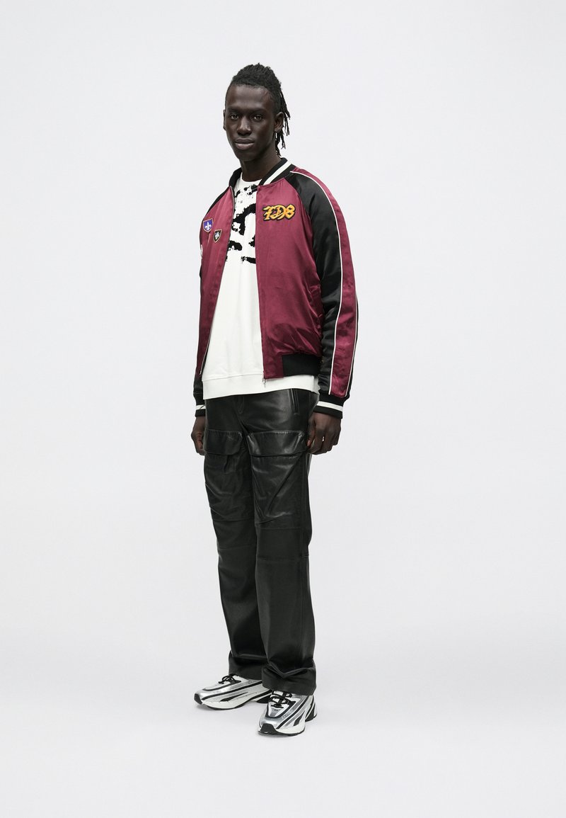 Tumepruun satiinist pommitaja-tüüp jakk plaastritega, valge graafilise mustriga pusa, mustad nahast cargo-püksid ning must-valged kingad.