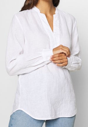 Kvinde iført en langærmet hvid hørbluse med mandarin-krave og blå jeans, der holder hænderne foldet i taljehøjde.