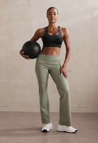 Sujetador deportivo negro, pantalones anchos de color verde oliva y zapatillas deportivas blancas. El modelo sostiene un balón de baloncesto negro. Texturas suaves y un buen ajuste.
