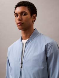 Giacca bomber azzurro chiaro realizzata in un materiale leggero e liscio. Presenta una chiusura con zip, colletto a coste e un logo sottile sul petto.