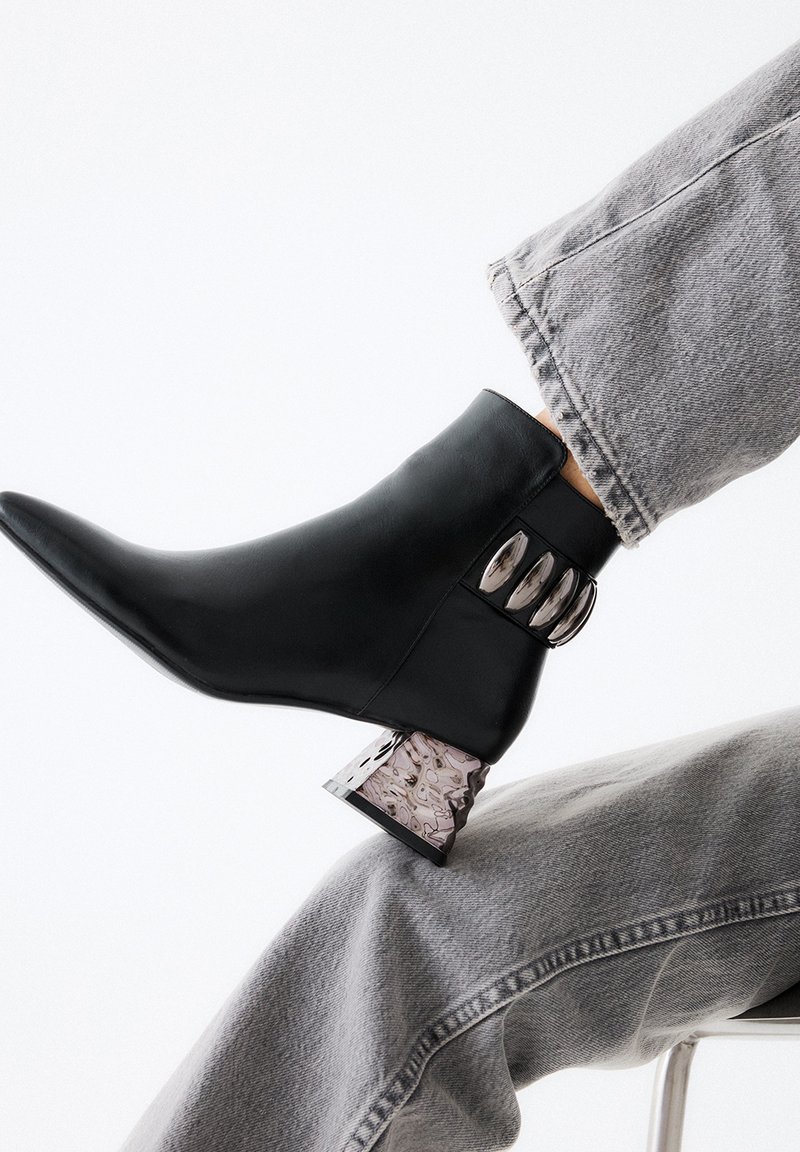 Botín negro de cuero con punta afilada que presenta un acento metálico en el tobillo y un exclusivo tacón grueso veteado. Lleva puesto con jeans gris claro.