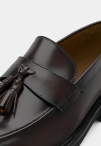 Mocassin en cuir marron avec une finition brillante, doté d'une bride et de détails à glands. La chaussure présente une forme élégante et des coutures discrètes.