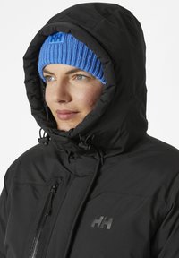 Veste isolante noire avec col montant et capuche réglable. Bonnet en tricot côtelé bleu avec logo. Texture lisse, zips fonctionnels.
