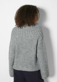 Personne aux cheveux bouclés portant un pull en tricot gris épais et un pantalon foncé, tournée de dos sur un fond blanc uni.