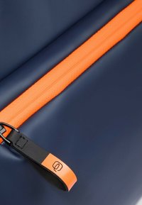 Bolsa azul marino con una cremallera naranja prominente y una lengüeta de cuero negro con un logotipo estampado. Material liso y duradero.