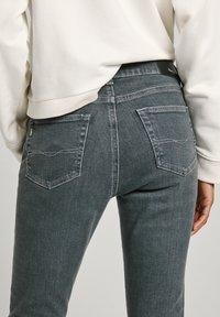 Jeans oscuros ajustados de color gris, con bolsillos traseros con solapas angulares y detalles de costura en contraste. Combinados con un sudadera blanca.