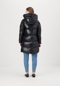Zwarte pufferjas met een capuchon, glanzende textuur, gestikte ontwerp en lange lengte. Gecombineerd met lichtblauwe jeans en zwarte schoenen.