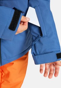Main tenant une carte blanche en train de la glisser dans la poche zippée de la manche d'une veste bleue, avec un pantalon orange visible en bas.