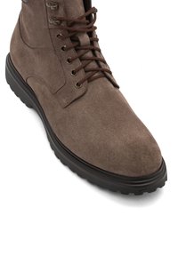 Botas de ante marrón con cordones y suela de goma negra, ojales de metal y ganchos para los cordones sobre un fondo blanco.