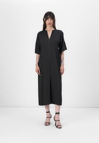 Black midi dress with short sleeves, deep V-neck, and front zipper. Smooth fabric, relaxed fit, and side slit, paired with black strappy heels.