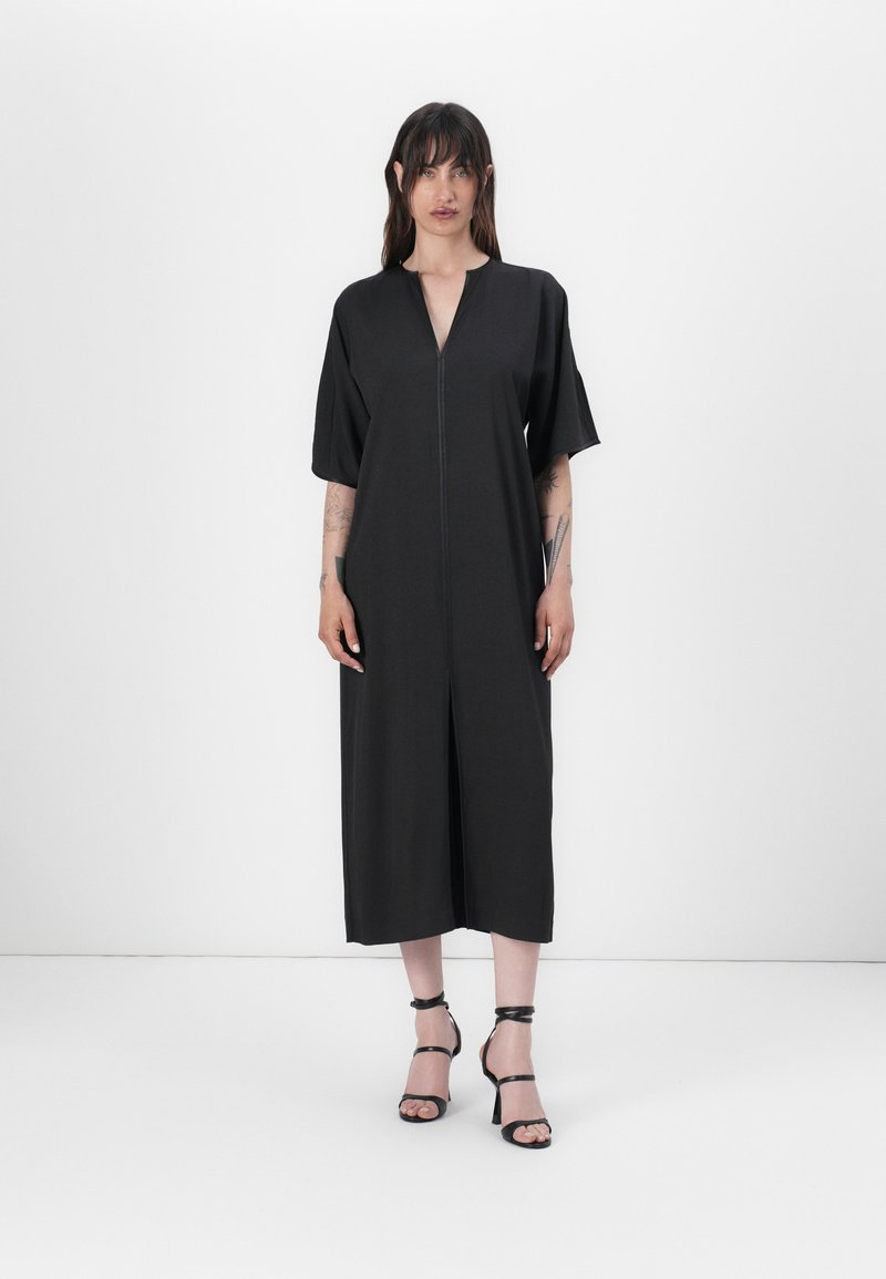 Black midi dress with short sleeves, deep V-neck, and front zipper. Smooth fabric, relaxed fit, and side slit, paired with black strappy heels.