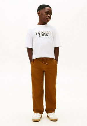 Camiseta blanca de manga corta con el texto "TOMMY 1985", combinada con pantalones marrones de cintura elástica y zapatillas blancas con suelas beige.