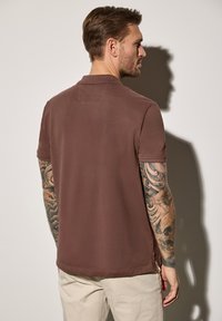 Polo marrón de manga corta, que cuenta con un collar, aberturas laterales y una textura suave. El modelo tiene los brazos tatuados y pantalones de color claro.