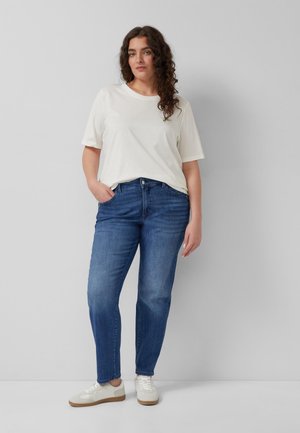 Frau mit lockigem braunem Haar, die ein weißes T-Shirt, blaue Jeans und weiße Turnschuhe trägt, steht mit einer Hand in der Tasche vor einer schlichten Wand.