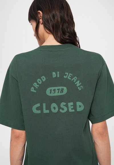 Πράσινο βαμβακερό T-shirt με κοντά μανίκια, που φέρει μεγάλο τύπωμα στην πλάτη: «CLOSED» και «PROD. DI JEANS 1978» σε έντονα, αντίθετα γράμματα.