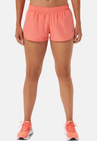 Mujer vestida con pantalones cortos deportivos color coral y zapatillas de running a juego, de pie con las piernas visibles desde la cintura hasta los pies contra un fondo blanco.