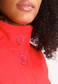 Manteau rouge en mélange de laine avec un col montant, doté de deux boutons ronds ornés d'accent argenté. Texture lisse avec une coupe ajustée.