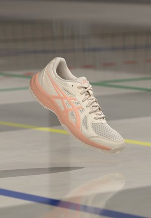 Zapatilla deportiva de color beige claro y melocotón con cordones, flotando sobre el suelo de una pista cubierta marcado con líneas de colores.