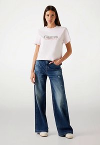 Tricou roz scurt cu un logo argintiu, asortat cu blugi largi albaștri, având zone estompate și detalii uzate, purtat cu adidași albi.