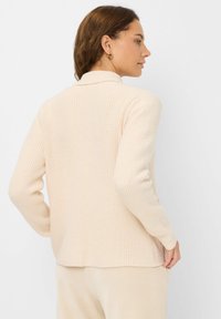Beige geribbelde cardigan met een kraag en lange mouwen, met een gestructureerde stof en een rechte zoom. Geen zichtbare sluitingen.